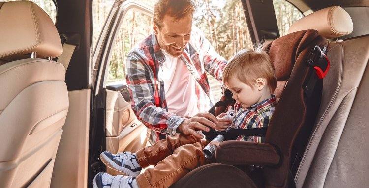 Father is adjusting child's car seat, while kid is playing with toy binoculars. Side view