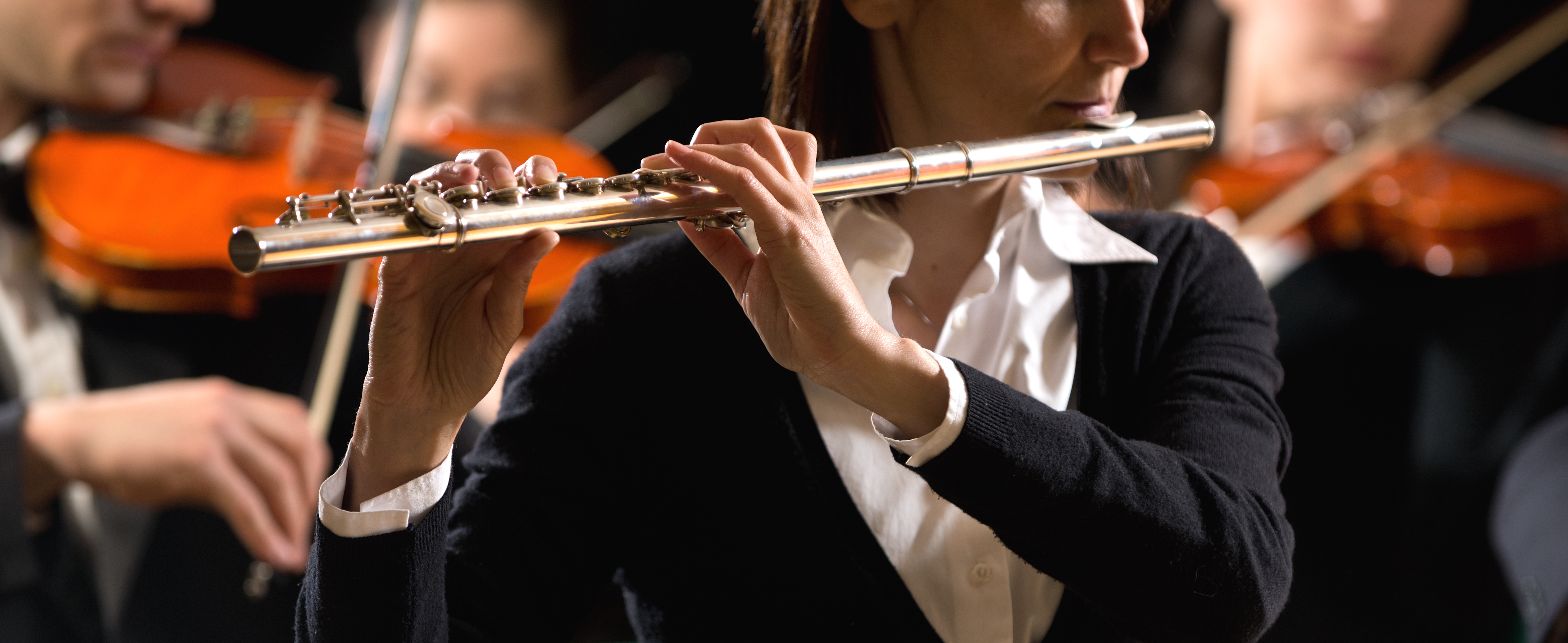 Professional female flutist in concert with symphony orchestra players on background.