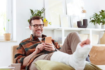 Pleased mature man wearing checkered shirt sitting with broken leg on sofa in living room at home and using smart phone.