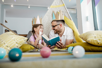 Father and his little girl are on the floor surrounded by toys, he is reading a story to his girl