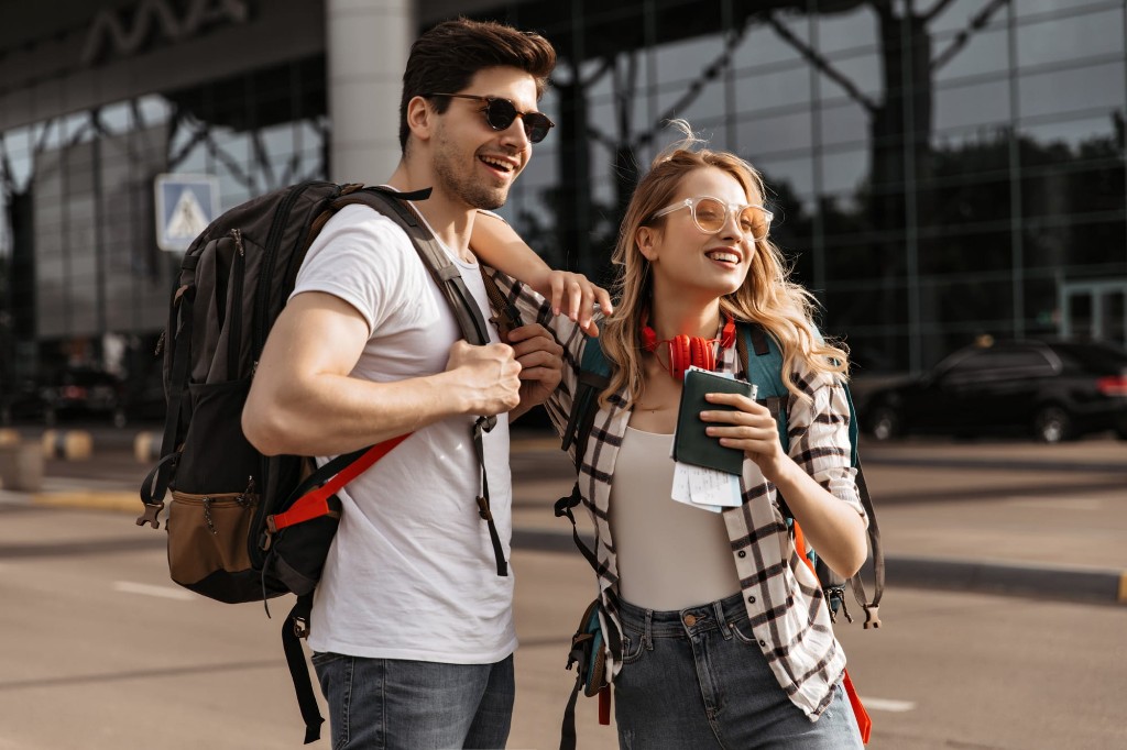 Pareja joven sonriente con mochilas, pasaporte y billetes frente a un aeropuerto, listos para viajar.