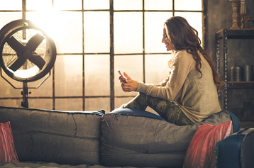 Seen from the side,a brunette woman is smiling, looking down at her phone sitting on the back of a sofa. Industrial chic ambiance and cozy atmosphere, sunlight is streaming through the loft window.; Shutterstock ID 278626649; PO: xxxxxxx; Job: xxxxxx; Client: xxxxxx; Other: xxxxx